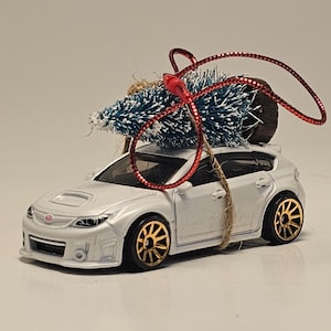 May include: A white toy car with gold wheels and a small blue Christmas tree tied to the roof with red ribbon. The car has a Subaru logo and is a hatchback model. A festive holiday ornament.