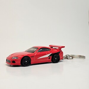 May include: Red toy car with a white stripe and black accents. The car has a rear spoiler and a keychain attached.