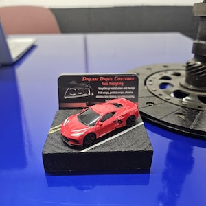 May include: A red toy car on a black base with a business card that reads "Dream Drive Customs Auto Restyling" with the text "Vinyl Wrap Installation and Design" and a list of services offered.