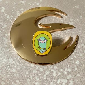 Könnte beinhalten: Eine goldfarbene Metallstifte mit einem grünen und gelben Emaille-Design. Das Design zeigt einen Cartoon-Vogel mit einem gelben Schnabel und weißen Augen. Der Vogel sitzt auf einem Ast.