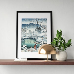 May include: A black framed print of a Japanese woodblock print depicting a snowy landscape with a bridge over a river. A small gold lamp and a potted plant are on a wooden shelf below the print.