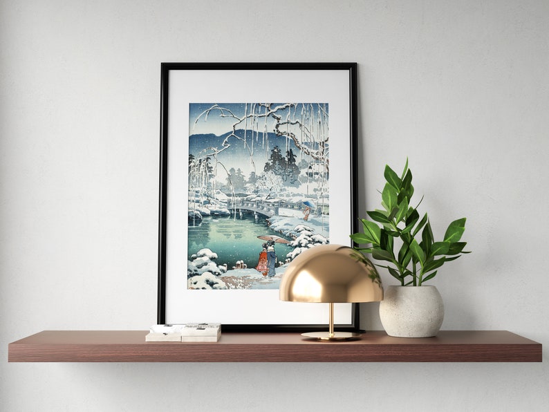 May include: A black framed print of a Japanese woodblock print depicting a snowy landscape with a bridge over a river. A small gold lamp and a potted plant are on a wooden shelf below the print.