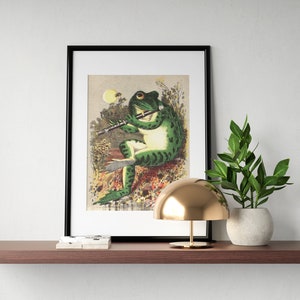 K&ouml;nnte beinhalten: Ein gerahmter Druck eines gr&uuml;nen Frosches, der in einer Waldlandschaft Fl&ouml;te spielt. Der Frosch sitzt auf einem Stein und ist von gr&uuml;nen Bl&auml;ttern und Blumen umgeben. Der Druck ist in einem schwarzen Rahmen und hat einen wei&szlig;en Passepartout.