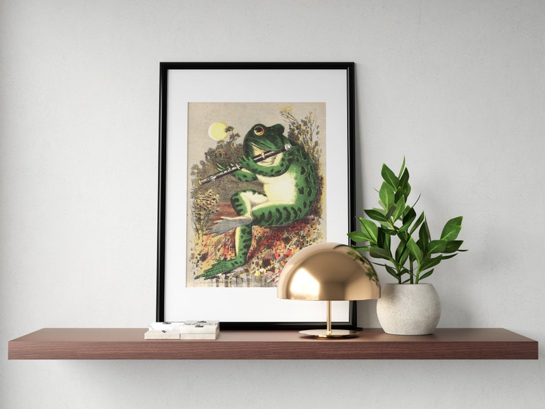 K&ouml;nnte beinhalten: Ein gerahmter Druck eines gr&uuml;nen Frosches, der in einer Waldlandschaft Fl&ouml;te spielt. Der Frosch sitzt auf einem Stein und ist von gr&uuml;nen Bl&auml;ttern und Blumen umgeben. Der Druck ist in einem schwarzen Rahmen und hat einen wei&szlig;en Passepartout.