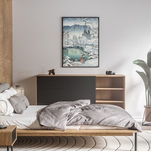 May include: A bedroom with a wooden bed frame, a grey duvet, and a black bedside table with a lamp. A black cabinet with shelves is in the background, and a potted plant is on the right side of the image. A framed print of a Japanese landscape scene hangs on the wall.