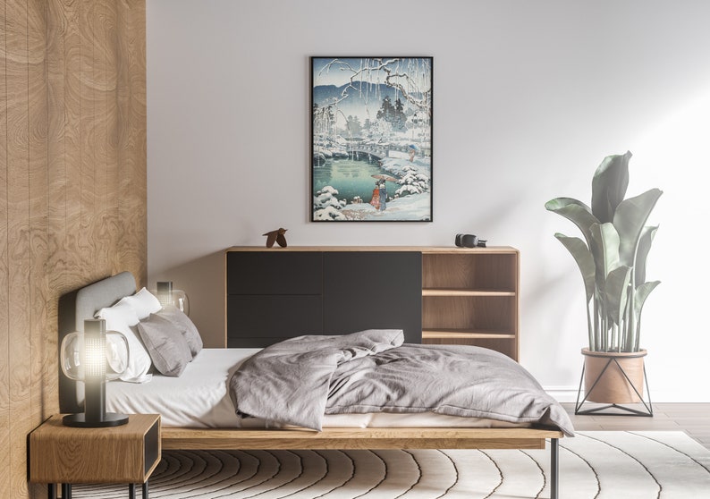 May include: A bedroom with a wooden bed frame, a grey duvet, and a black bedside table with a lamp. A black cabinet with shelves is in the background, and a potted plant is on the right side of the image. A framed print of a Japanese landscape scene hangs on the wall.