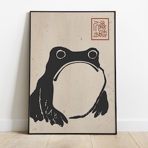 Peut inclure: Illustration en noir et blanc d'une grenouille sur un fond beige avec un tampon rouge qui dit "福" en caractères japonais.