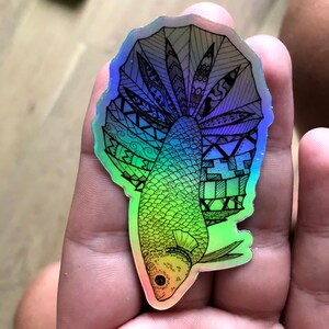 Puede incluir: Una pegatina hologr&aacute;fica con un dibujo en blanco y negro de un pez con una aleta grande y detallada. El pez est&aacute; delineado en negro y la aleta est&aacute; llena de patrones intrincados. La pegatina tiene un acabado iridiscente arco&iacute;ris.