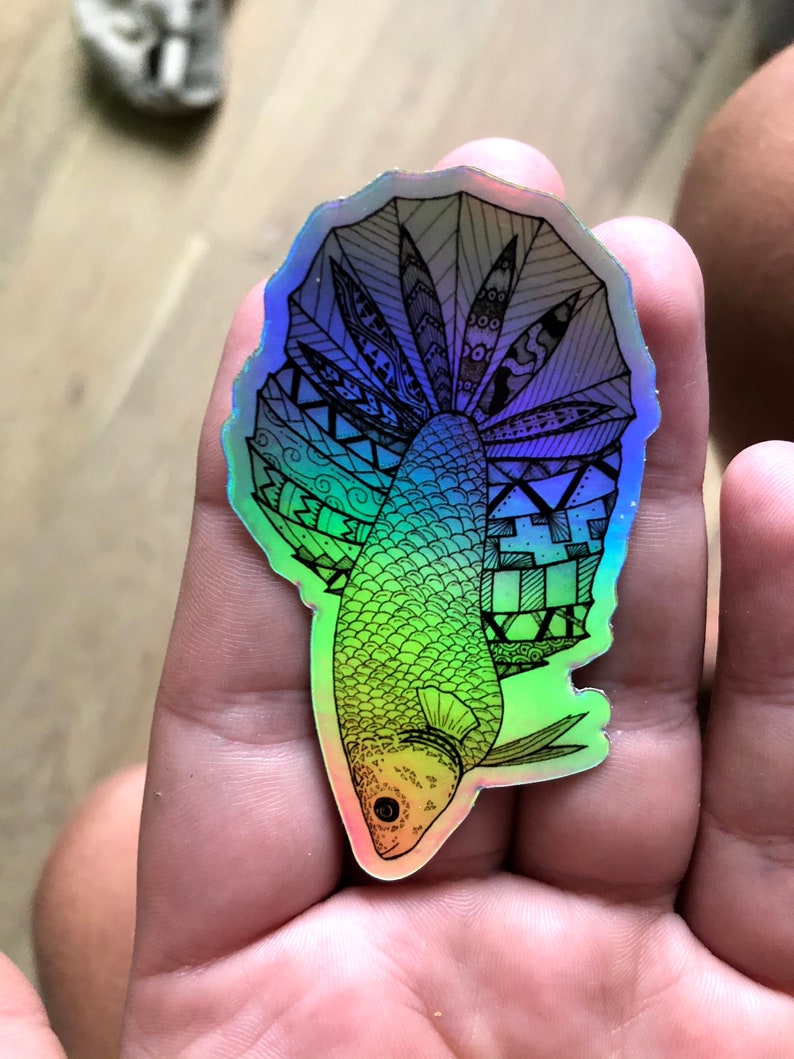Puede incluir: Una pegatina hologr&aacute;fica con un dibujo en blanco y negro de un pez con una aleta grande y detallada. El pez est&aacute; delineado en negro y la aleta est&aacute; llena de patrones intrincados. La pegatina tiene un acabado iridiscente arco&iacute;ris.