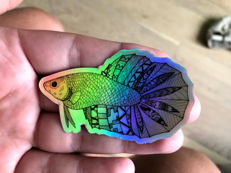 Puede incluir: Una pegatina hologr&aacute;fica con un dibujo detallado en l&iacute;nea negra de un pez betta sobre un fondo arco&iacute;ris iridiscente.