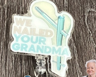 May include: A clear plastic badge holder with a blue and white design that says "We Nailed Your Grandma". The badge holder has a retractable cord and a blue heart charm with a smiley face. The badge holder is on a wooden surface.