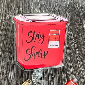 May include: Red plastic sharps container with the text "Stay Sharp" on the front. A gold-colored syringe charm is attached to a clear plastic badge holder with a red glitter button. A red syringe with a red blood drop is in the background. The text "V-spin Designs Creative Crafts" is at the bottom of the image.