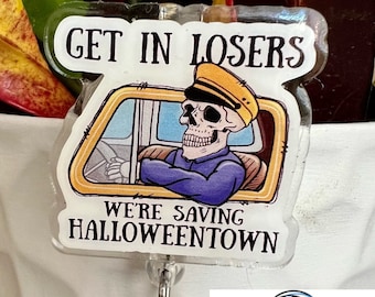 May include: A Halloween-themed badge reel with a skeleton driver in a taxi, the text "GET IN LOSERS WE'RE SAVING HALLOWEENTOWN", and a charm of a smiling snowman wearing a top hat. The reel is attached to a white pot with green and red plants.