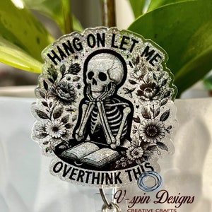 May include: A silver glitter badge holder with a black and white skeleton holding a book. The badge holder has the text "Hang on let me overthink this" and a black smiley face charm.