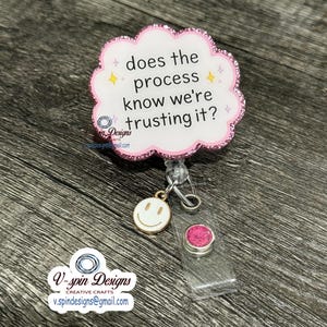 May include: A cloud-shaped badge reel with a pink glitter border and white center featuring the black text "does the process know we're trusting it?" accented by small yellow and pink stars. A gold smiley face charm and a pink glitter button hang below.