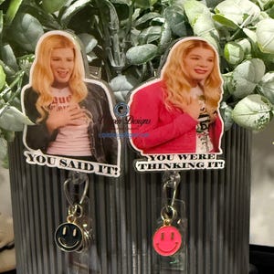 May include: Two badge reels featuring images of a blonde woman with the phrases "YOU SAID IT!" and "YOU WERE THINKING IT!". Each reel has a smiley face charm. One charm is black and gold, the other is pink.