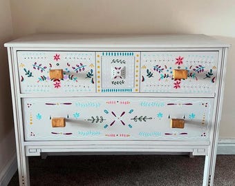 May include: A light gray wooden chest of drawers featuring two levels. The top level has two drawers and a central square panel, while the bottom level has one wide drawer. All surfaces are decorated with hand-painted floral and leaf motifs.