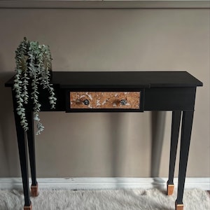 May include: A black console table with a single drawer featuring a cork-like pattern. The table has four legs with gold accents and is sitting on a white fluffy rug.