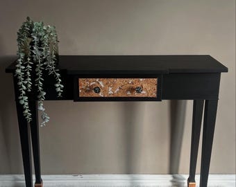 May include: A black console table with a single drawer featuring a cork-like pattern. The table has four legs with gold accents and is sitting on a white fluffy rug.