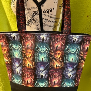 May include: A black tote bag with a repeating pattern of a horror movie scene. The scene features a house with a figure holding a knife and a figure with a mask. The bag has black straps.