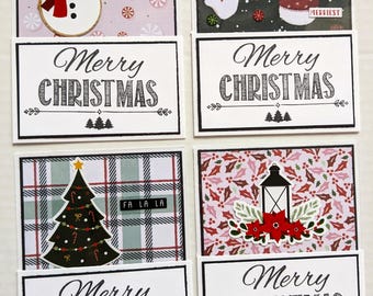 May include: Four rectangular greeting cards featuring festive holiday designs. Each card displays a snowman, snow people, a Christmas tree, or a lantern with holly. Below each image, a white card section reads Merry Christmas in black text.