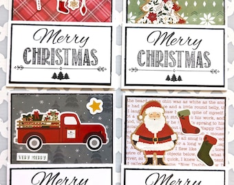 May include: Four rectangular Christmas greeting cards featuring festive designs including a red plaid pattern with pine branches, a green patterned tree with the word BELIEVE, a red truck carrying gifts, and a Santa Claus figure with stockings.