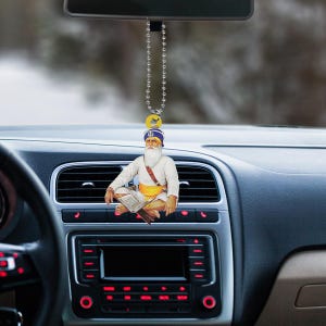 May include: A car hanging ornament featuring a seated figure with a white beard, turban, and traditional attire. The ornament is suspended from a silver chain and hangs in front of a car dashboard. The figure is holding a book.