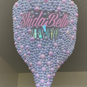 May include: A white plastic hair brush with a pink and iridescent design. The brush is covered in small, iridescent beads and has the text "Shida Belle" in pink lettering. The brush also has a graphic of a hair clipper, comb, and scissors.
