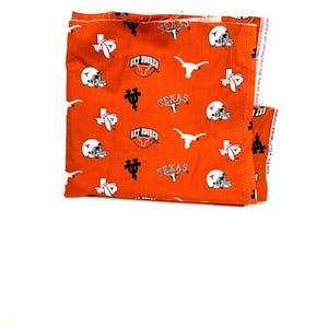 May include: An orange fabric with a repeating pattern of white football helmets, the Texas Longhorns logo, and the word "TEXAS". The fabric is folded and appears to be new.