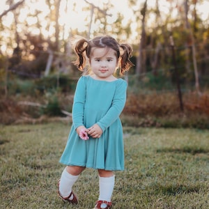 May include: A young girl with pigtails wears a teal blue long-sleeved dress and white socks. She is holding a pink flower and standing in a grassy field.