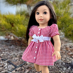 Könnte beinhalten: Eine Puppe mit langen schwarzen Haaren, die ein rot-weiß kariertes Kleid mit einer weißen Schleife trägt. Die Puppe steht auf einem Bett aus Holzspänen.