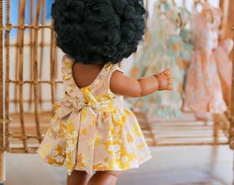 May include: A doll wearing a yellow and pink floral dress with a bow in the back. The doll has dark curly hair and is standing in front of a wicker cage.