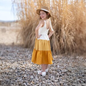 May include: A tiered dress with a white bodice, a yellow midsection, and a mustard-colored skirt. The dress is paired with a straw hat with a white ribbon and white sandals. The dress is worn by a person standing on a bed of rocks.