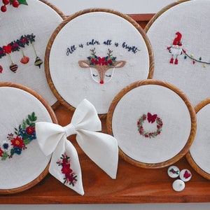 May include: Six embroidery hoops with various Christmas-themed designs, including a reindeer with the text "All is calm, all is bright", a wreath, a garland, two gnomes holding a string of lights, and a floral arrangement. A white bow is in the center of the image.