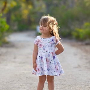 May include: A young girl wearing a white dress with a floral print stands on a dirt path. The dress has pink and purple flowers on it. The girl is looking to the right of the frame.