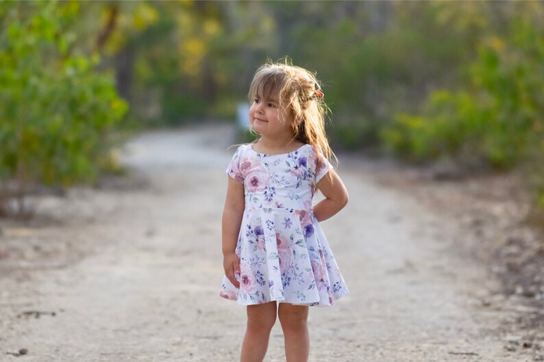 May include: A young girl wearing a white dress with a floral print stands on a dirt path. The dress has pink and purple flowers on it. The girl is looking to the right of the frame.
