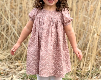 May include: A young girl wearing a pink floral dress with white puff sleeves and white tights. She is standing in a field of tall brown grass.