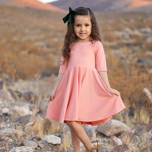 May include: A young girl wearing a pink dress with a green velvet bow in her hair. She is standing in a rocky desert landscape.