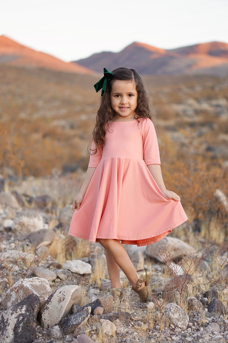 May include: A young girl wearing a pink dress with a green velvet bow in her hair. She is standing in a rocky desert landscape.