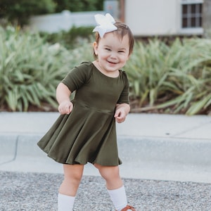 May include: A young child wearing a green dress and white socks smiles while standing on a sidewalk.