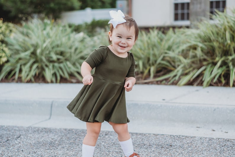 May include: A young child wearing a green dress and white socks smiles while standing on a sidewalk.