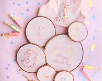 May include: Pink and white embroidery hoops with various designs including balloons, a number three, a cupcake, and the words "Happy Birthday".