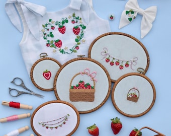 May include: A set of embroidery hoops featuring various strawberry designs, including a basket of strawberries, a garland of strawberries, and a single strawberry. There are also spools of thread, scissors, and a small white linen dress with a strawberry embroidery design.