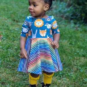 May include: A young girl wearing a blue and rainbow striped dress with a pattern of animal faces. She is wearing yellow knee-high socks and black boots.