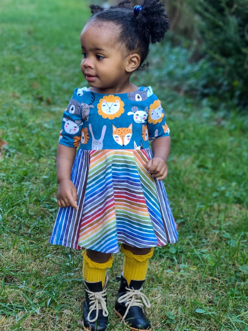 May include: A young girl wearing a blue and rainbow striped dress with a pattern of animal faces. She is wearing yellow knee-high socks and black boots.