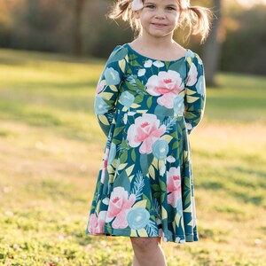 May include: A young girl wearing a teal blue dress with a floral print. The dress has long sleeves and a flared skirt. She is wearing brown ankle boots and has a pink bow in her hair.