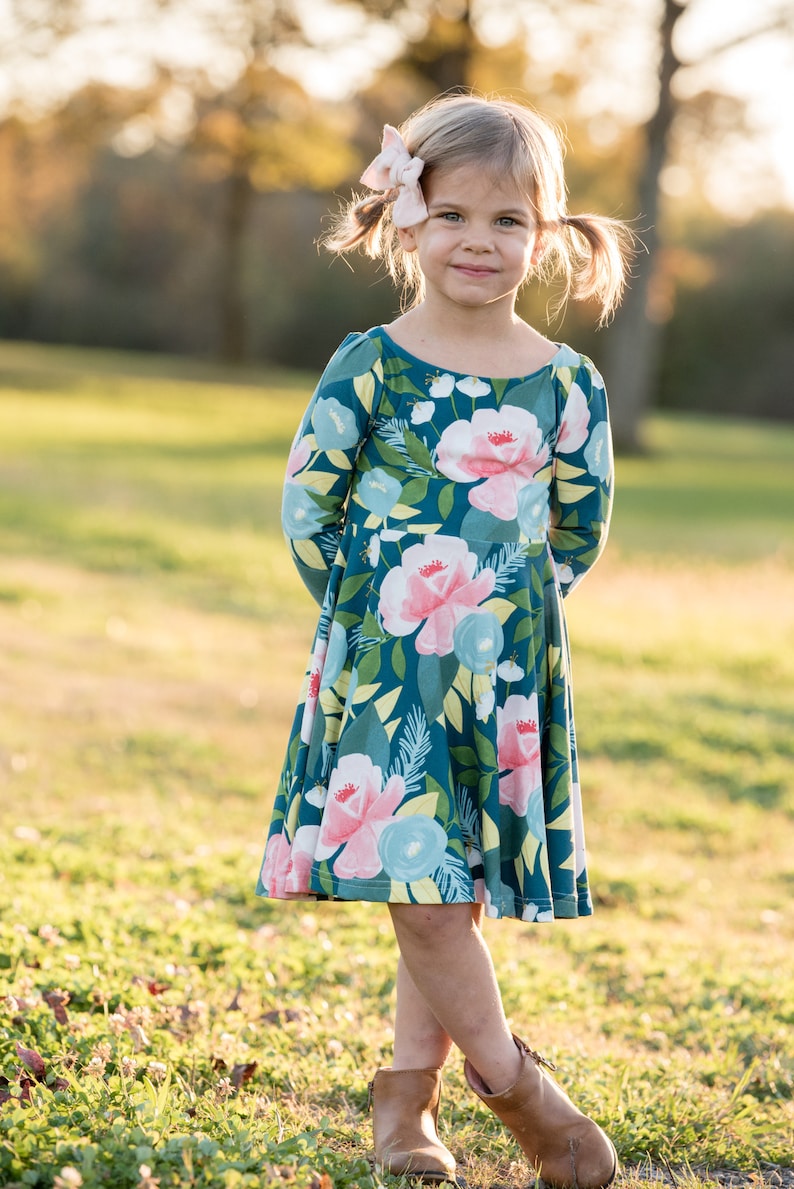 May include: A young girl wearing a teal blue dress with a floral print. The dress has long sleeves and a flared skirt. She is wearing brown ankle boots and has a pink bow in her hair.