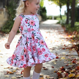 May include: A young girl wearing a white dress with a pink floral pattern and a navy blue bow in her hair. She is wearing white socks and black shoes. She is standing on a sidewalk with fallen leaves.