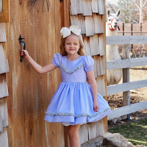 May include: A young girl in a light blue dress with a white bow in her hair stands in front of a wooden door. She is holding the door handle with her left hand and looking at the camera.