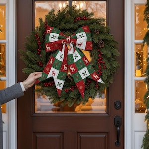 Może przedstawiać: Wieniec bożonarodzeniowy z dużą, dekoracyjną kokardą. Kokarda ma czerwony, zielony i biały wzór z motywami reniferów i płatków śniegu. Wieniec wykonany jest z gałęzi iglastych i czerwonych jagód.